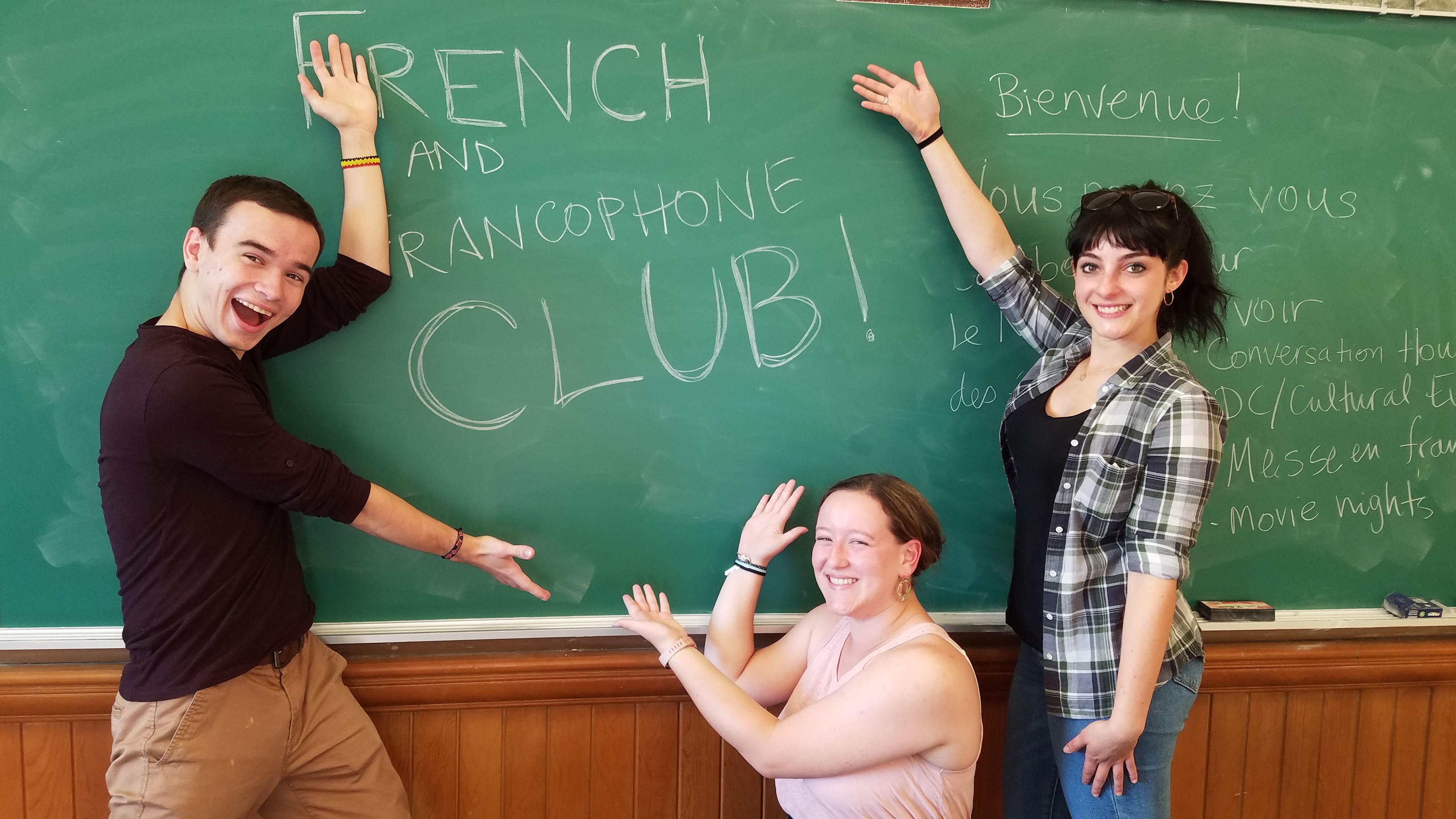 Smiling students pointing at a French Club sign.
